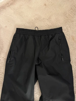 Svarta täckbyxor med dragkedjor - Svarta täckbyxor med elastisk midja och raka ben. Byxorna har två praktiska fickor med dragkedjor framtill och är tillverkade i ett vattenavvisande material, perfekta för kalla dagar utomhus. Snygg och enkel design som funkar till vinterns aktiviteter.