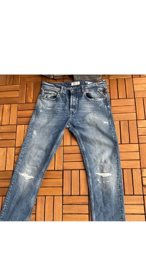 Replay Anbass blå jeans med slitningar - Snygga Replay Anbass jeans i klassisk blå tvätt med coola slitningar och detaljer på både fram- och baksida. Modellen har raka ben och normal passform, med fem fickor och broderade bakfickor. Perfekt för dig som gillar en avslappnad streetstil.