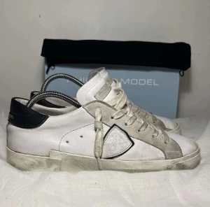 Philippe Model  - Säljer ett Philippe Model sneakers i vitt skinn med grå mockadetaljer och svart häl. Klassisk låg modell med rund tå, snörning och platt sula. Skorna har en cool sköldlogga på sidan och en stilren vibe som gör din outfit snyggare! Skostorleken är 40 och kartong medföljer! Kan skicka via post. Skriv om ni är intresserade!