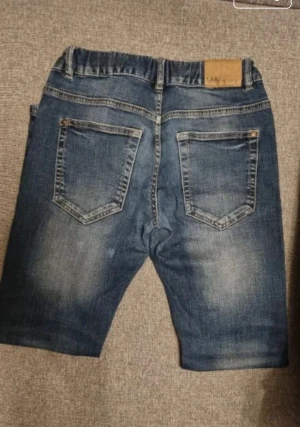 Blå jeans från LAB Industries - Snygga blå jeansshorts från LAB Industries med klassisk femficksdesign och elastisk midja. Shortsen har en normal passform och är tillverkade i bomull med slitningar för en cool look. Perfekta för sommarens alla äventyr.