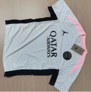 PSG fotbollströja och shorts Jordan - Säljer ett PSG matchställ från Jordan med vit och svart kortärmad tröja, rosa detaljer på axlarna och svarta shorts med rosa inslag. Tröjan har Qatar Airways-tryck och PSG-logga, tillverkad i lätt polyester som andas. Shorts med elastisk midja och PSG/Jordan-loggor.