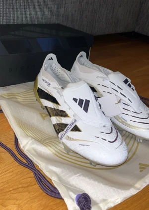 Adidas Predator fotbollsskor vit/guld - Adidas Predator fotbollsskor i vit med svarta och guldiga detaljer. Snygg design med täckt snörning och Predator-logga på sidan. Skorna har en modern form och är tillverkade i syntetmaterial med Hybridtouch-teknologi för extra komfort och kontroll på planen. Helt o andvända