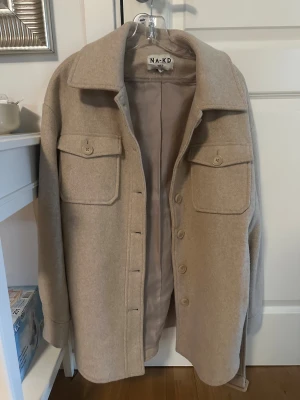 Beige jacka från NA-KD - Snygg beige  jacka från NA-KD med stora bröstfickor, knappar framtill och bälte i midjan. Jackan har klassisk krage och är långärmad, perfekt för lager-på-lager. Materialet är mjukt och känns lyxigt, med en clean och stilren look.