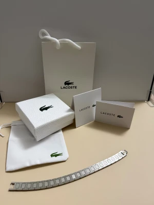 Lacoste armband i rostfritt stål - Snyggt armband från Lacoste i silverfärgat rostfritt stål med rektangulära länkar och graverade Lacoste-loggor. Kommer med original ask, påse och certifikat. Perfekt accessoar för dig som gillar stilrena detaljer och ikonisk design.
