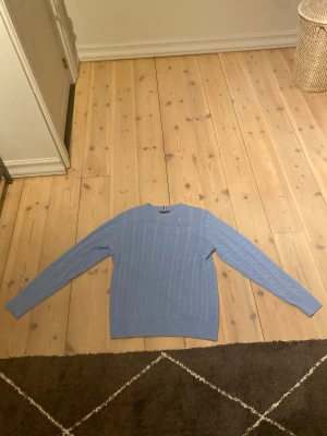 Ljusblå kabelstickad tröja Tommy Hilfiger - Snygg ljusblå kabelstickad tröja från Tommy Hilfiger med rund hals och diskret logga på bröstet. Tröjan har långa ärmar och ribbade muddar vid hals, ärmslut och nederkant. Perfekt för dig som gillar klassisk stil med en modern touch. Den är oanvänd. 