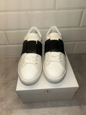 Valentino Garavani Open sneakers vit/svart - Snygga Valentino Garavani Open sneakers i vitt skinn med bred svart läderdetalj över vristen. Klassisk rund tå, vita snören och ikoniska nitar baktill på sulan. Perfekt för dig som vill ha en clean men lyxig look.