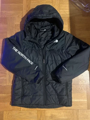 Svart pufferjacka från The North Face - Svart pufferjacka från The North Face med huva och dragkedja. Jackan har logga på bröstet och ärmen, samt diskreta reflexdetaljer. Tillverkad i syntetmaterial och har en quiltad look. Perfekt för kyliga dagar och riktigt snygg streetstil.