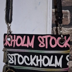 Svart axelväska med rosa text- Robin Ruth  - Robin Ruth -Cool svart axelväska med rosa och vit graffiti-aktig text där det står 'STOCKHOLM' över hela väskan. Väskan har silvriga metalldetaljer, dragkedja och avtagbar axelrem. 