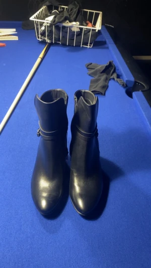 Svarta stövletter från ECCO, storlek 40 -  OBS SKORNA HAR SLDRIG ANVÄNDS UTOMHUS. Mamma tyckte klackarna var för höga. Snygga svarta stövletter från ECCO i storlek 40. Skorna har rund tå, hög klack och dragkedja på sidan. Tillverkade i skinnimitation med slät finish och diskret detalj runt ankeln. Perfekt för dig som gillar stilrena boots med lite höjd.