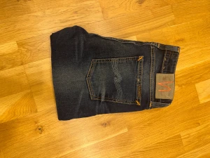 Nudie Jeans blå 29/30 - Snygga mörkblå jeans från Nudie Jeans i modellen skinny med klassiska orangea sömmar och coola slitningar. Jeansen har fem fickor, normal passform och är tillverkade i mjukt bomullsmaterial. Perfekt för dig som gillar en smal siluett och stilren look.
