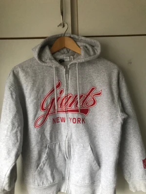 Grå Giants hoodie med dragkedja - Säljer en grå hoodie från  med röd logga och texten 'New York' på bröstet. Tröjan har huva med snörning, framfickor och dragkedja. Perfekt för dig som gillar sportig stil och vill ha något bekvämt och snyggt.