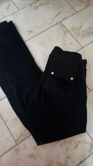 Svarta mjukisbyxor Juicy Couture XS - Svarta mjukisbyxor från Juicy Couture i storlek XS. Byxorna har en bred ficka med silverfärgade knappar och snörning i midjan med metalländar. Mjukt material 