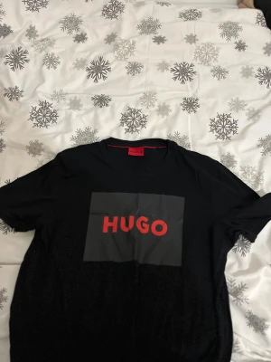 Svart HUGO t-shirt med röd logga - Snygg svart t-shirt från HUGO med stor röd logga på bröstet. Klassisk rund hals och korta ärmar. T-shirten har en stilren design med kontrasterande tryck framtill. Perfekt för dig som gillar streetwear och vill sticka ut med ett känt märke.