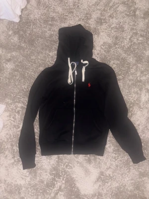 Svart hoodie från Polo Ralph Lauren - Svart hoodie från Polo Ralph Lauren med vit snörning och dragkedja framtill. Klassisk röd Polo-logga broderad på bröstet. Tröjan har huva, långa ärmar och två fickor framtill. Perfekt för chill dagar och streetwear-stil. Funkar både till tjej och kille 