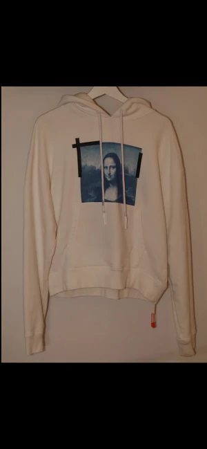 Äkta Off-White vit hoodie ”Monalisa” - Säljer en clean vit hoodie från Off-White med ett stort blått konsttryck på bröstet ”monalisa” och svarta detaljer. Hoodien har snörning med Off-White-logga och en röd tag vid nederkanten. Mjuk bomullskvalitet och klassisk känguruficka framtill. Taggen går att scanna för att se att den är äkta. Nypris: 6678kr