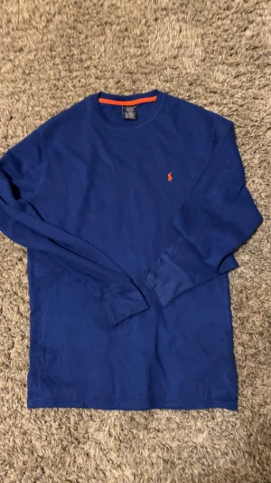 Blå långärmad tröja från Polo Ralph Lauren - Blå långärmad tröja från Polo Ralph Lauren med orange logobrodyr på bröstet och en orange detalj vid halsringningen. Tillverkad i 100% bomull, perfekt för dig som gillar klassisk stil med en färgklick. Enkel och snygg design som funkar året runt.