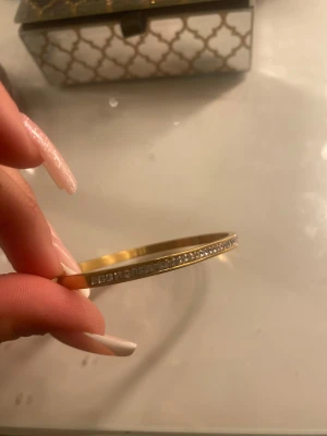 Guldfärgat armband med stenar - Stilrent guldfärgat armband med raka kanter och en rad infattade, fyrkantiga klara stenar längs ovansidan. Armbandet är stelt och öppet i ena änden, vilket gör det enkelt att ta på och av. Perfekt för dig som gillar en clean och glittrig look.