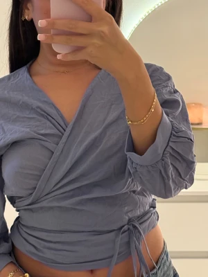 Blå omlottopp med knyt i midjan - Säljer en blå omlottopp med snygg v-ringning och knyt i midjan. Toppen har trekvartsärm med rynkade detaljer som ger en cool look. Perfekt att styla med jeans eller kjol för en trendig vibe.