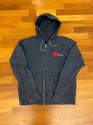 Mörkblå hoodie från Replay S - Snygg mörkblå hoodie från Replay med dragkedja och stor huva. Framsidan har ett broderat rött R och Replay-logga i vitt. Mjuk insida och klassisk känguruficka. Perfekt för chill dagar och streetwear-stil.