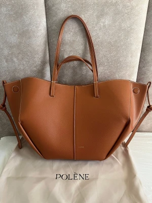 Polène brun handväska i skinn - Säljer en snygg handväska från Polène i brunt skinn med unik geometrisk form. Väskan har två långa handtag, vita kontrastsömmar och magnetlås. Insidan är rymlig med en innerficka och ljus textil. Perfekt för dig som gillar stilrena accessoarer.