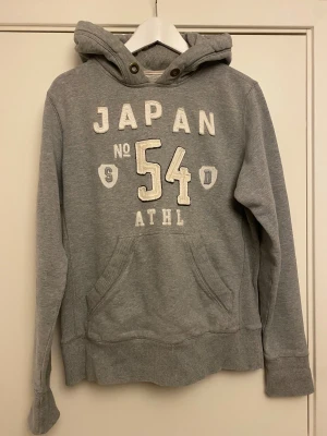 Grå hoodie Superdry Japan Spirit M - Snygg grå hoodie från Superdry i storlek medium med broderad text 'JAPAN 54 ATHL' på bröstet och Superdry-logga i huvan. Klassisk känguruficka framtill och ribbade muddar. Mjuk bomullsblandning, perfekt för chill dagar.