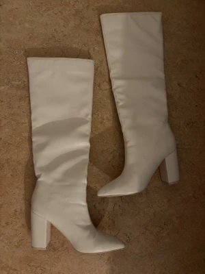 Vita höga boots med klack - Säljer ett par stilrena vita boots med hög klack och spetsig tå. Skorna är knähöga och har en snygg, minimalistisk design som verkligen sticker ut. Perfekta för dig som vill ha ett statement piece i garderoben. Materialet är slätt och ger en lyxig känsla.
