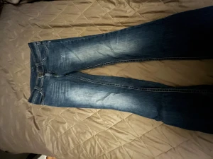 Blå bootcut jeans med broderade fickor - Snygga blå jeans med bootcut passform och coola broderade detaljer på bakfickorna. Jeansen har slitningar och en liten reva under ena bakfickan( bild 3) Klassisk femficksmodell med ljusare tvätt på låren och bak. Perfekt för dig som gillar retrovibe och avslappnad stil. 