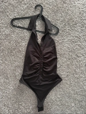 Svart omlott bodysuit med knappar - Snygg svart bodysuit med omlottkrage och knappar framtill. Modellen är ärmlös och har halterneck med bred krage. Materialet är mjukt och stretchigt, troligen polyester. Perfekt för att skapa en cool och trendig look.