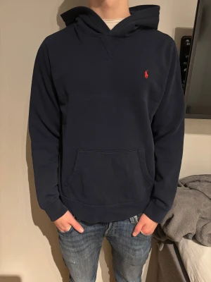 Mörkblå hoodie från Polo Ralph Lauren - Snygg mörkblå hoodie från Polo Ralph Lauren med röd broderad logga på bröstet. Klassisk känguruficka framtill och skön huva. Mjuk bomullsmix som är perfekt för chill dagar. Enkel och stilren design som funkar till jeans eller joggers.