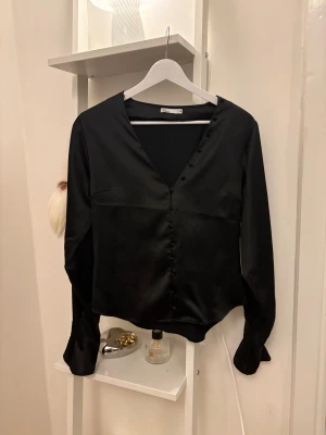 Svart satinblus - Svart blus i satin med v-ringning och knappar framtill. Blusen har långa ärmar med lätt utsvängda ärmar längst ner 🤍