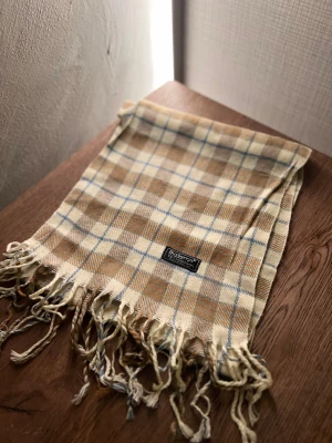 Rutig ullhalsduk från Burberrys - Klassisk rutig halsduk från Burberrys i 100% ull. Halsduken har fransar i ändarna och den ikoniska Burberrys-etiketten. Perfekt accessoar för att lyfta din outfit. Anledningen att de stör ”burberrys” är för att de är en äldre modell.