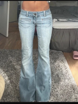 Bootcut jeans från Dollhouse, ljusblå - Snygga ljusblå bootcut jeans från Dollhouse med låg midja och klassiska fem fickor. Jeansen har slitna detaljer, knapp och dragkedja framtill samt broderad logga i gult. Perfekt för dig som gillar retrovibe och breda ben.ena knappen på bakfickan finns ej på men inget som man tänker på