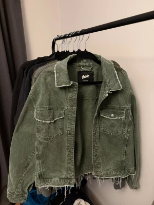 Grön jeansjacka från Pull&Bear S - Snygg grön jeansjacka från Pull&Bear med råa kanter och fransad finish. Jackan har två bröstfickor med lock och knappar, klassisk krage och avslappnad passform. Perfekt för dig som vill ha en cool och avslappnad stil.