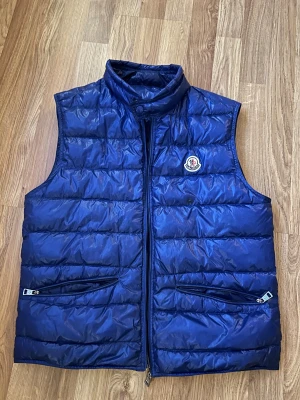 Blå dunväst från Moncler Longue Saison - Snygg blå dunväst från Moncler Longue Saison med hög krage och två dragkedjefickor framtill. Västen har quiltad design och klassisk Moncler-logga på bröstet. Perfekt för lager-på-lager och ger en clean look till din outfit.