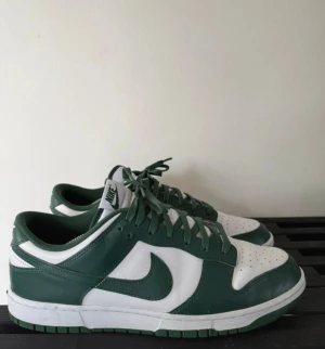 Nike Dunk Low gröna och vita sneakers - Nike Dunk Low sneakers i grönt och vitt med klassisk Swoosh på sidan. Skorna har snörning, platt sula och rund tå. Ovandelen är i skinn och färgblocken ger en clean och sportig vibe. Perfekta för dig som gillar streetwear och vill sticka ut med snygga färger.  Använda 3 gånger 