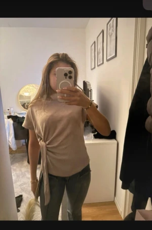 Beige kortärmad topp med knyt - Säljer en beige kortärmad topp med snygg knytning i sidan. Toppen har en enkel och clean look med rund halsringning och är tillverkad i ett mjukt material som känns skönt mot huden. Perfekt att styla med jeans eller kjol.