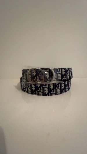  bälte med monogram och silverdetalj - Snyggt bälte  med klassiskt svart och grått omonogrammönster och en stor silvrig spänne framtill. Bältet har en bred design och är perfekt för att lyfta vilken outfit som helst med en lyxig touch. Materialet är textil med tydlig logga  över hela bältet.