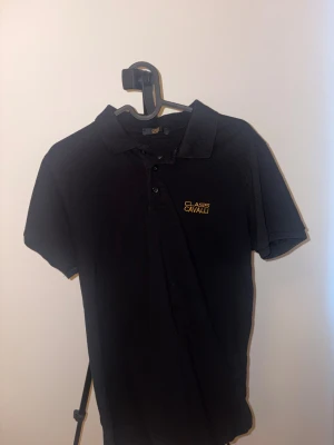 Svart pikétröja Class Cavalli - Svart pikétröja från Class Cavalli med gul broderad logga på bröstet. Klassisk krage och tre knappar framtill. Kortärmad och tillverkad i mjuk bomull som är skön mot huden. Perfekt för dig som gillar stilrena och enkla plagg med en touch av lyx.