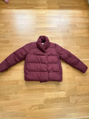 Vinröd pufferjacka med krage - Säljer en vinröd pufferjacka med bred krage och långa ärmar. Jackan har ett quiltat mönster och stängs med knappar på insidan. Perfekt för dig som vill ha en varm och snygg jacka med lite oversized känsla. Köpt för 700, knappt använd, säljer för 430