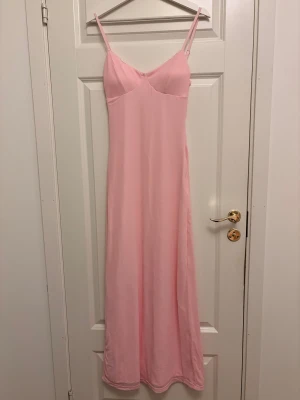 Ljusrosa långklänning med smala axelband - Supersöt ljusrosa långklänning med smala justerbara axelband och mjuk, slät yta. Klänningen har en feminin v-ringad överdel med sömmar som ger fin passform. Materialet känns lätt och följsamt, perfekt för varma dagar. Dragkedja i sidan för enkel påklädning.