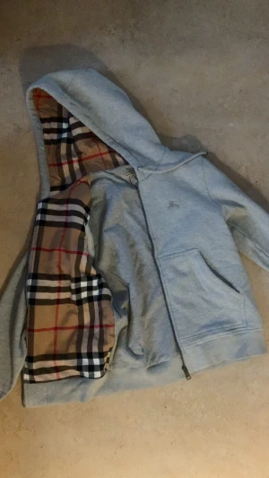 Grå Burberry Kofta - Grå kofta från Burberry med klassiskt rutig insida i beige, svart, vit och röd. Jackan har dragkedja framtill, huva med snörning och stora fickor. Tillverkad i 100% bomull, perfekt för dig som gillar stilrena streetwear-vibes och exklusiva detaljer.