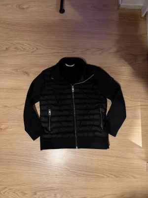 Svart cardigen från Moncler - Svart cardigen från Moncler med quiltad framsida och två dragkedjeförsedda fickor. Jackan har långärmade släta ärmar och hel dragkedja framtill. Materialet är en mix av dun och syntet, perfekt för dig som gillar stilren och sportig look.