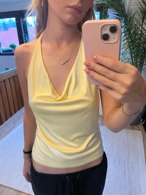 Ljusgul halterneck linnetopp - Snygg ljusgul halterneck topp med draperad v-ringning och öppen rygg. Tunn och glansig, perfekt för varma dagar. Sitter tight och har en croppad passform som ger en fräsch och trendig look.