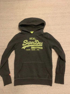 Mörkgrå Superdry hoodie med tryck - Snygg mörkgrå hoodie från Superdry med stort gult tryck på bröstet och japanska tecken. Tröjan har huva, magficka och långa ärmar med ribbade muddar. Insidan är mjuk fleece för extra komfort. Perfekt för chill dagar och streetstyle.