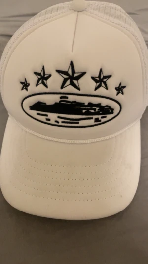 Vit Corteiz truckerkeps med mesh - Säljer en vit truckerkeps från Corteiz med svart broderad logga och stjärnor framtill. Kepsen har mesh-paneler på sidorna och bak, samt broderad Corteiz-text baktill. Märkesband på insidan och gul etikett. Perfekt för dig som vill sticka ut.