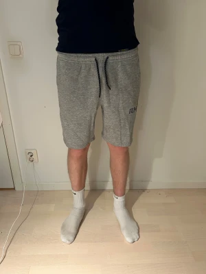 Grå mjukisshorts med snörning RM - Grå mjukisshorts från RM med svart snörning i midjan och diskret logga på vänster ben. Shortsen har normal passform, sidofickor och är tillverkade i mjukt bomullsmaterial. Perfekta för chill eller träning, riktigt sköna och stilrena.