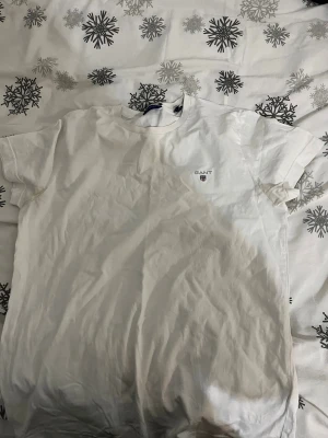 Vit t-shirt från GANT med logga - Snygg vit t-shirt från GANT med klassisk rund hals och liten broderad logga på bröstet. T-shirten är kortärmad och gjord i mjuk bomull, perfekt för en clean och enkel stil. Passar dig som gillar stilrena plagg med diskret branding.