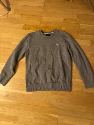 Grå sweatshirt från Henri Lloyd - Snygg grå sweatshirt från Henri Lloyd med rund hals och broderad logga på bröstet. Tröjan har ribbade muddar vid ärmslut och nederkant, samt en klassisk passform. Perfekt för chill dagar och enkel att matcha med jeans eller joggers. Storlek L men sitter som en M