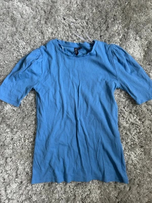 Blå ribbad topp från Pieces XS - Säljer en ljusblå ribbad topp från Pieces i storlek XS. Toppen har korta puffärmar och rund halsringning. Materialet är mjukt och stretchigt, perfekt för en chill och snygg look. Passformen är normal och toppen har en enkel, stilren design.