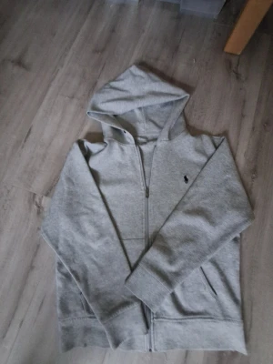 Grå hoodie från Polo Ralph Lauren - Snygg grå hoodie från Polo Ralph Lauren med klassisk broderad logga på bröstet. Tröjan har huva, dragkedja framtill och två fickor. Tillverkad i mjuk bomullsmix, perfekt för chill dagar. Långärmad och med ribbade muddar för extra komfort.